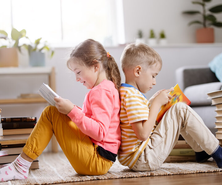 Zwei Kinder sitzen Rücken an Rücken und schauen sich ein Kinderbuch an